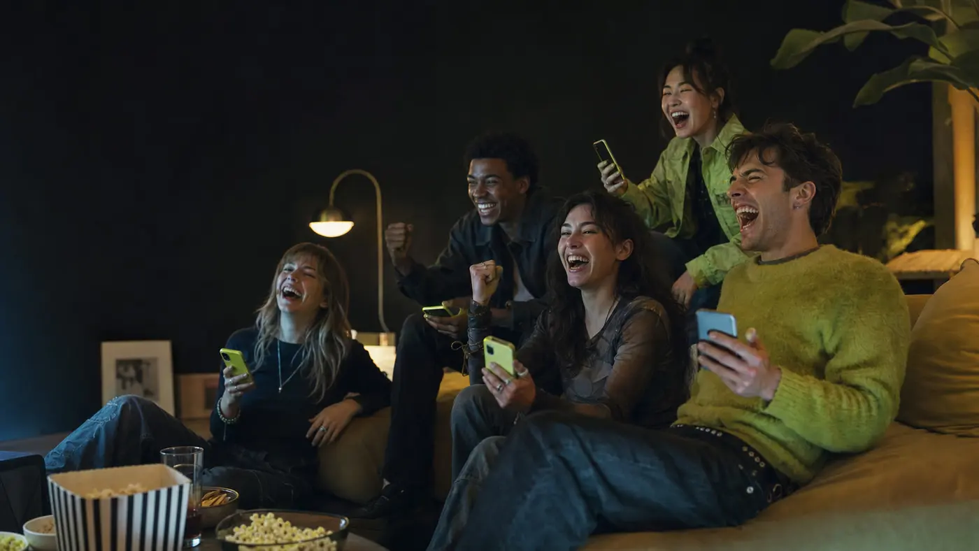 Une bande d'amis rit et réagit ensemble devant la télé, chacun son téléphone en main sur wazap